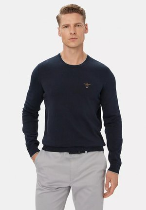 Giovane uomo con capelli biondi corti che indossa un maglione blu navy e pantaloni grigio chiaro, in piedi con una mano in tasca.