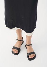 Sandali in pelle nera con suola piatta, cinturino alla caviglia regolabile, fascia larga per le dita dei piedi e punta squadrata. Texture liscia e design minimalista.