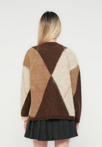 Frau mit langen blonden Haaren, die einen flauschigen, geometrisch gemusterten Pullover in Braun, Beige, Creme und einen plissierten Rock trägt und sich von einem weißen Hintergrund abwendet.