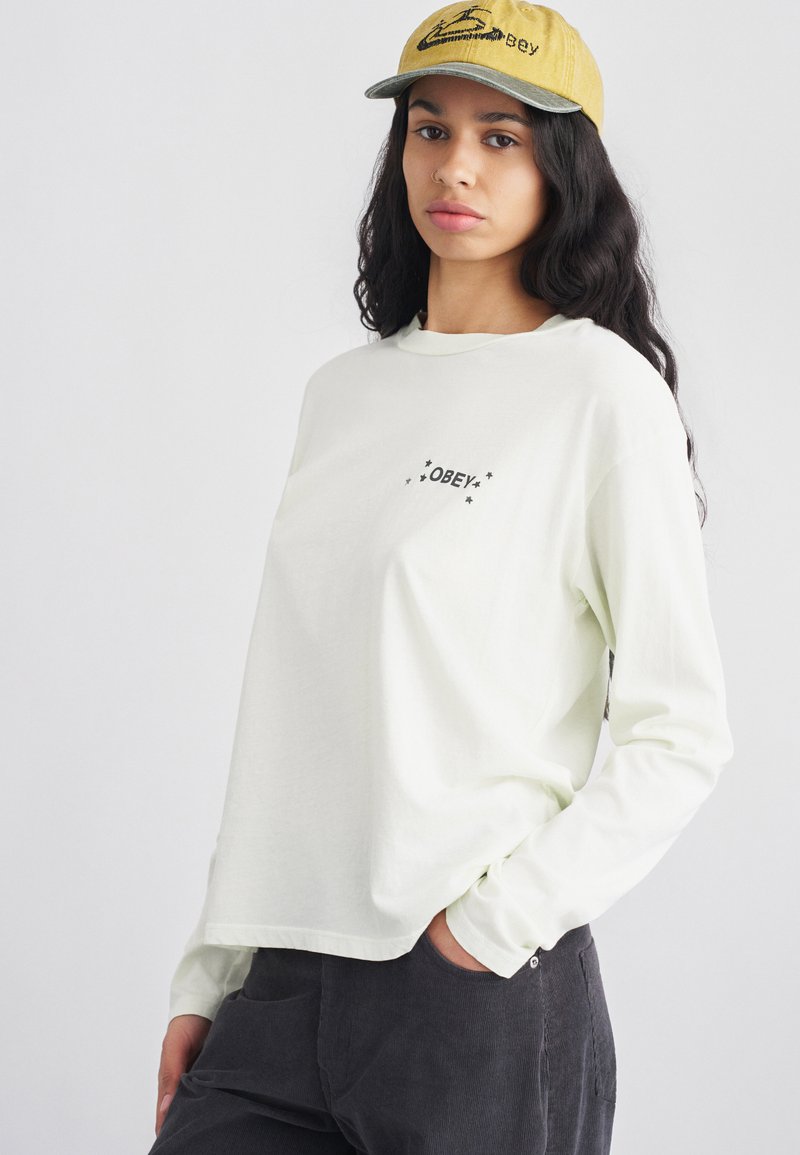 Lichtgroen shirt met lange mouwen en "OBEY"-logo in het zwart met sterren, gecombineerd met een gele pet met geborduurde details.