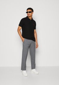 Schwarzes Polo-Shirt mit kurzen Ärmeln und rotem Logo, kombiniert mit hellgrauen Hosen und weißen Sneakers, vor einem einfarbigen Hintergrund.