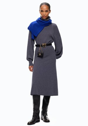 Donna che indossa un vestito a collo alto grigio in maglia, cintura nera con fibbia dorata e mini borsa, sciarpa blu drappeggiata su una spalla e stivali neri al ginocchio.