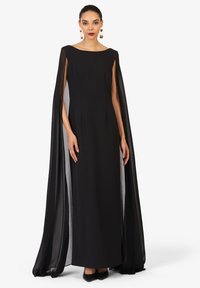 Zwarte avondjurk met een vloeiende cape, voorzien van een bootnek en zijsplitten. Soepele textuur, vloerlengte, gecombineerd met puntige zwarte hakken.