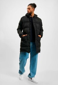 Jachetă puffer neagră cu glugă, având un design matlasat, închidere frontală cu nasturi și buzunare laterale. Purtați peste un top închis la culoare și blugi de un albastru deschis.