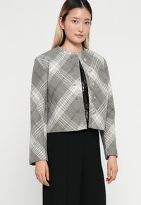 Giacca cropped a righe grigie con maniche lunghe, caratterizzata da un collo arrotondato e chiusure con bottoni in argento, indossata sopra un top nero.