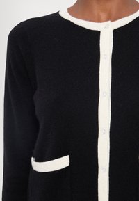 Black knitted cardigan with white trim, featuring a buttoned front and a small patch pocket on the left side. Textured fabric.