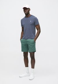 Stripete marineblå og hvit t-skjorte, grønne shorts, hvite sneakers og en lys rosa caps. Modellen står med hendene i lommene mot en ensfarget bakgrunn.