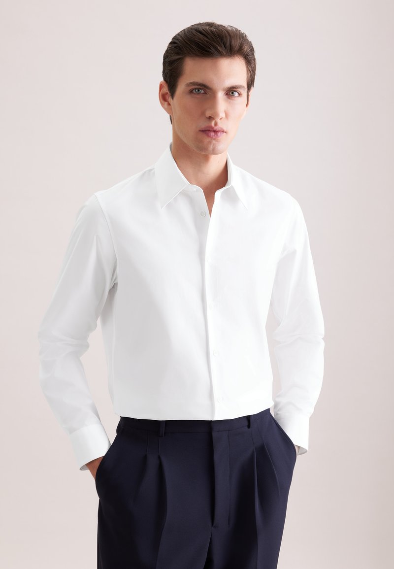 Giovane uomo con capelli corti e scuri che indossa una camicia bianca abbottonata e pantaloni plissettati blu scuro, mani nelle tasche, sfondo neutro.