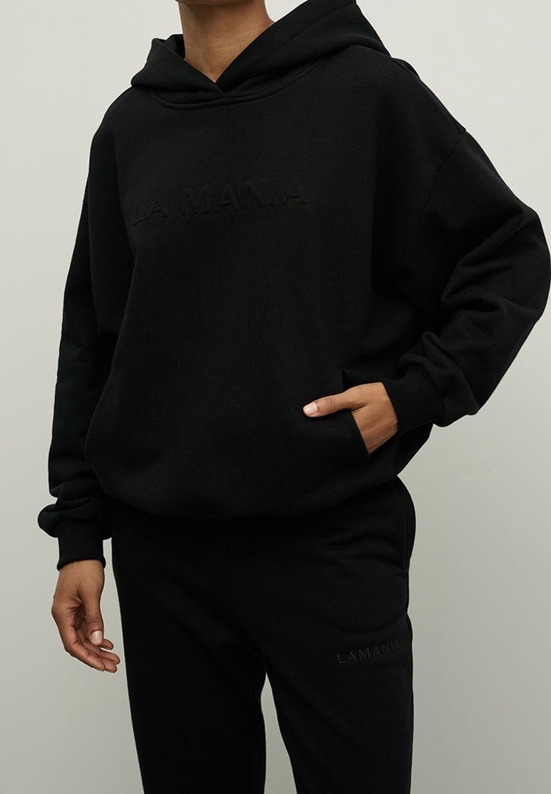 Personne portant un sweat à capuche noir et un pantalon noir assorti avec un texte en relief subtil "LAMANIA" sur les deux, une main dans la poche, sur fond uni.
