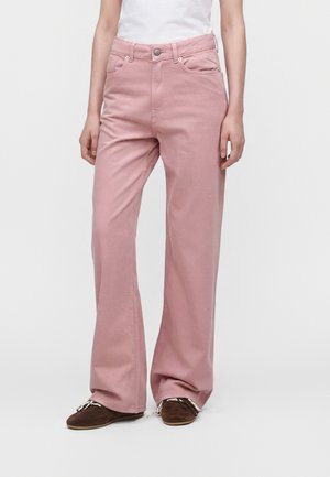 Femme portant un pantalon rose taille haute à jambes larges avec poches avant et chaussures marron à lacets, debout devant un fond blanc uni.
