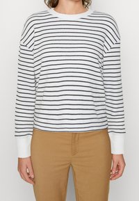 Maglione bianco a coste con righe orizzontali navy, scollo ampio e maniche lunghe. Abbinato a pantaloni marrone chiaro.