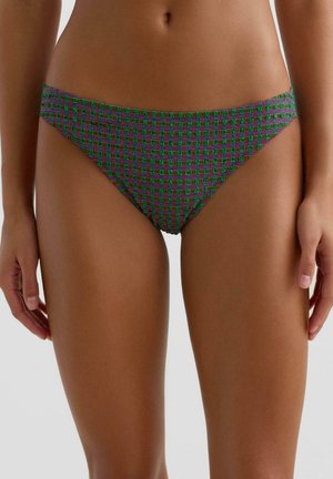 Kobieta w zielono-fioletowych bikini w kratę, stojąca z rękami swobodnie opuszczonymi wzdłuż ciała na jednolitym tle.