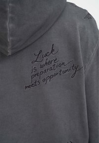 Sudadera gris con una textura suave, presenta un texto bordado en negro que dice: "La suerte es donde la preparación se encuentra con la oportunidad". Diseño simple.