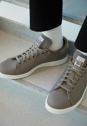 Personne portant des baskets Adidas Stan Smith grises avec des semelles blanches et des chaussettes blanches, debout sur des escaliers mouchetés.