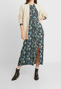 Blommig maxiklänning i mörk marinblå med gröna och blå mönster; har en hög slits framtill och är kombinerad med en beige, grovt stickad cardigan och bruna ankelskot. 