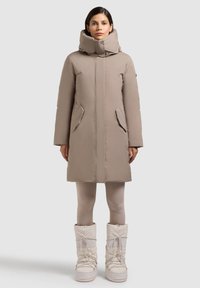 Béžový vatovaný kabát s vysokým, vyplneným golierom, predným zipsom a bočnými vreckami. Nosený s svetlo béžovými legínami a robustnými bielymi snehovými topánkami.