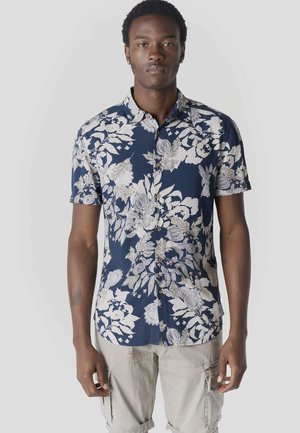 Hombre con camisa azul marino de manga corta con botones y estampado floral blanco, y pantalones cortos cargo beige, de pie contra un fondo liso.