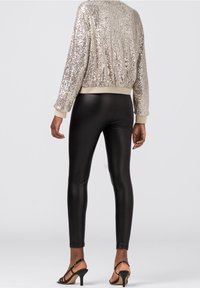 Glitzernde silberne Pailletten-Bomberjacke mit cremefarbenem Besatz, kombiniert mit engen schwarzen Leggings und schmalen schwarzen High Heels. Rückansicht, die die Textur hervorhebt.