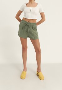 Wit cropped top met een vierkante halslijn en korte mouwen, gecombineerd met losse groene shorts met een elastische tailleband en een trekkoord. Gele klompen.