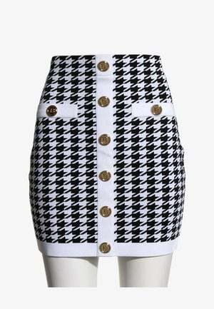 Mini-jupe pied-de-poule noir et blanc avec bordure blanche, boutons dorés au centre et deux pattes décoratives boutonnées aux hanches.