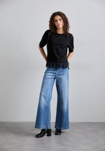 Fekete, rövidujjú felső csipkés díszítéssel, világoskék, bőszárú farmernadrággal párosítva. A modell fekete bokacsizmát visel, szürke háttér előtt áll.