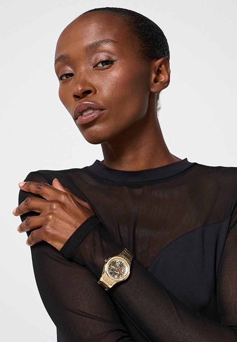 Reloj de pulsera dorado con una correa metálica y una esfera redonda, que presenta un dial oscuro y engranajes visibles, llevado en una mano de piel oscura.