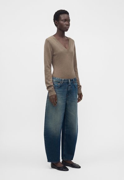 Femme debout portant un pull beige à col en V, un jean bleu délavé à jambes larges et des chaussures plates noires sur un fond clair uni.