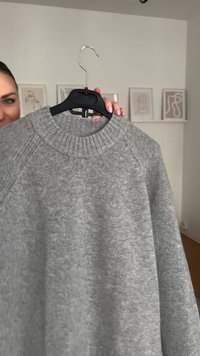 Harmaa neulepaita, jossa on ribbattu pyöreä pääntie, pehmeä rakenne ja raglan-hihat, roikkumassa mustassa muovihengarissa.