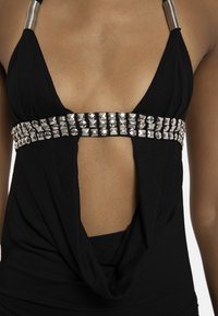 Top negru cu decolteu adânc în V, bandă lată argintie cu ținte sub bust și bretele argintii pe umeri, care arată zona abdomenului și a pieptului.