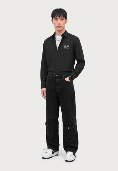 Jeune homme debout portant une chemise noire à manches longues, un pantalon cargo noir, des baskets blanches et un collier chaîne argenté sur un fond uni.