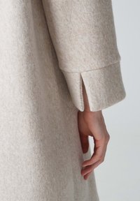 Manteau en laine beige avec une finition douce et texturée. Caractéristiques d'une coupe décontractée, fentes aux poignets et design simple et épuré. Main reposant sur le tissu.