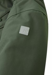 Tessuto di giacca verde con una superficie liscia, caratterizzato da una patchetta quadrata grigia sulla spalla e cuciture dettagliate.