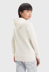 Sudadera de color crema con bolsillo canguro, capucha ajustable con cordón y puños acanalados, combinada con jeans de color azul claro.