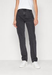 Vaqueros negros de talle alto confeccionados en denim, con un corte recto, bolsillos frontales y una sutil textura desgastada. Combinados con zapatillas blancas.