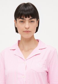 Rosa og hvit stripete pysjamasoverdel med korte ermer, krage med spiss, frontlomme og brodert design på lommen.