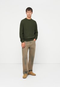 Maglione verde lavorato a maglia con maniche lunghe, abbinato a jeans beige chiaro a gamba dritta e sneakers color cammello. Design semplice, texture minima.