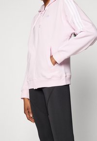 Růžová mikina na zip vyrobená z měkké látky, zdobená bílými pruhy na rukávech a malým logem na hrudi. V kombinaci s černými legínami.