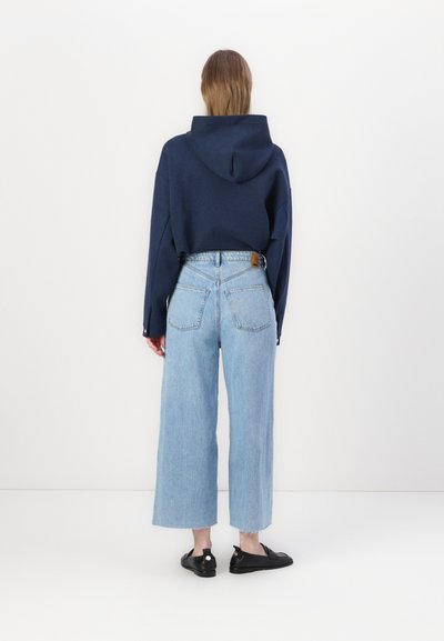 Sweatshirt à capuche bleu marine avec des manches larges, associé à un jean clair, taille haute et coupe large. Chaussures noires plates avec un accent blanc.