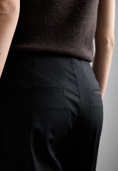Črne krojene hlače z gladko teksturo, ki imajo dva majhna zadnja žepa in eleganten dizajn, nosijo pa s temno rjavim zgornjim delom.