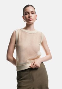 Ung kvinde med slicket hår iført guldøreringe, beige ærmeløs mesh-top og olivengrønne højtaljede bukser, poserende med hånden på hoften.