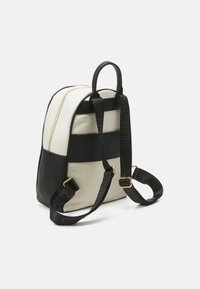 Mochila con una mezcla de lona y cuero, que presenta un cuerpo color crema, base negra, acentos de logo, cremallera dorada y correas ajustables.