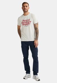 Creme t-shirt med røde "Petrol Outdoor Supply" tekst, parret med mørkeblå cargo bukser og hvide sneakers. Korte ærmer, afslappet pasform.