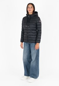 Zwarte pufferjack met capuchon, geborduurd ontwerp en ritssluiting; gedragen over losse blauwe jeans en witte sneakers.