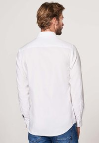 Weißes Hemd mit langen Ärmeln, das einen Standkragen, eine glatte Textur und einen geschwungenen Saum aufweist, kombiniert mit blauen Jeans.