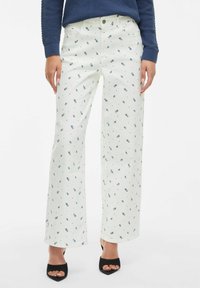Modèle portant un pantalon large blanc avec des motifs floraux bleus, associé à un pull bleu marine et des talons ouverts noirs.
