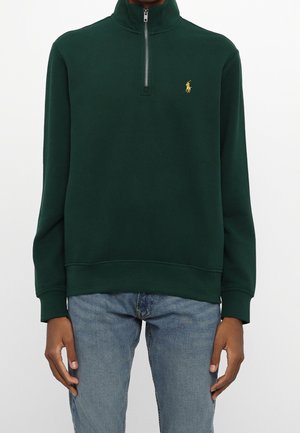 Dunkelgrüner Pullover mit Viertel-Reißverschluss und goldenem Pferdelogo auf der Brust, getragen zu hellblauen Jeans, Hände entspannt an den Seiten.