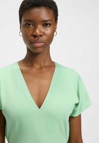 Vihreä bleiseri, jossa on syvä V-pääntie ja lyhyet hihat, sekä sileä, pehmeä rakenne. Hienovarainen ompelu pääntien ympärillä.