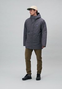 Veste isolée grise avec une fermeture éclair à l'avant, capuche réglable et poches latérales. Portée avec un pantalon kaki et des chaussures noires, debout sur un fond neutre.
