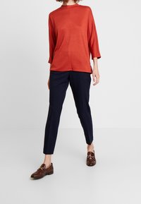 Rotes Langarmoberteil aus weichem Stoff, kombiniert mit marineblauen, schmal geschnittenen Hosen. Braune Slipper mit einer Schnallendetail. Einfaches, modernes Design.