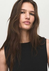 Model with long, wavy brown hair, wearing a black sleeveless top. Simple design with a round neckline and a smooth texture.